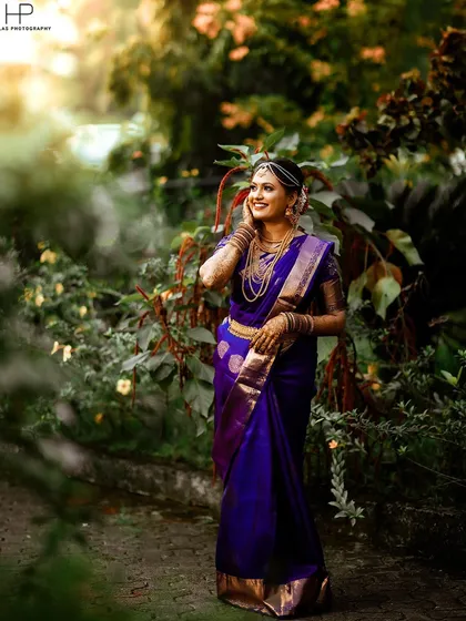 A stunning bridal portrait of the bride in her deep purple Kanjeevaram saree, set against a lush green backdrop. I used natural light to highlight the intricate gold jewelry and the joyful expression on her face.