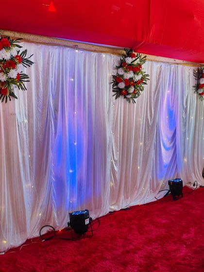 A simple and effective decor idea for a birthday or small function. A white curtain backdrop with fairy lights and blue uplighting, paired with a red carpet, instantly creates a dedicated photo or celebration corner.