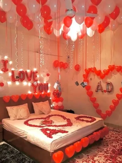 An elaborate romantic room decoration featuring ceiling balloons, an 'I LOVE YOU' light-up sign, and a large heart on the wall made of photos and fairy lights.