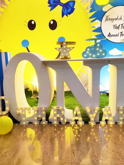 A close-up of a "First Trip Around the Sun" themed setup, showing the 'ONE' marquee table and 'THRAYAKSH' marquee letters.