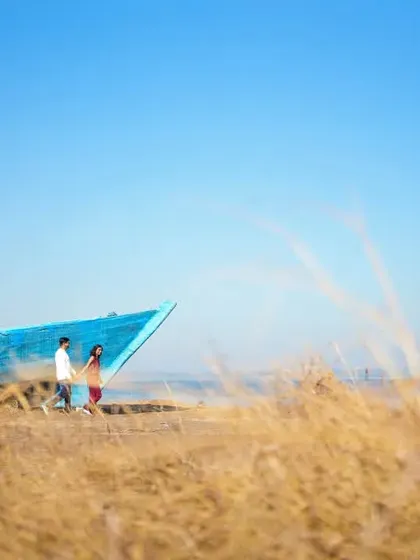 A wide shot taken through golden wild grass, framing a couple walking near an old boat. The vibrant blue of the sky and boat contrasts with the warm tones of the landscape for a visually striking image.