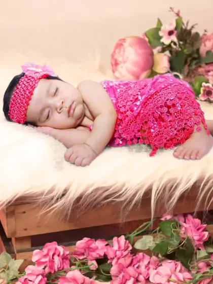 A newborn in a bright pink outfit sleeping on a wooden crate surrounded by pink flowers.