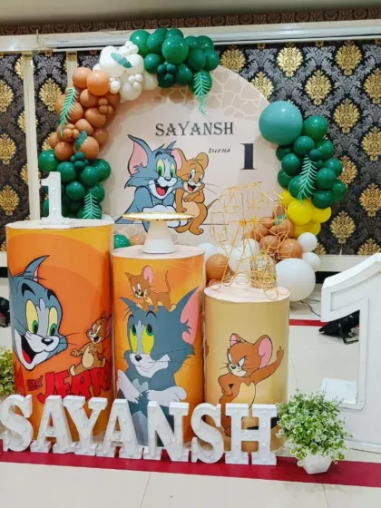 A Tom & Jerry first birthday with a jungle twist. The decor includes themed plinths, a green and brown balloon arch, and large marquee numbers for the big '1'.