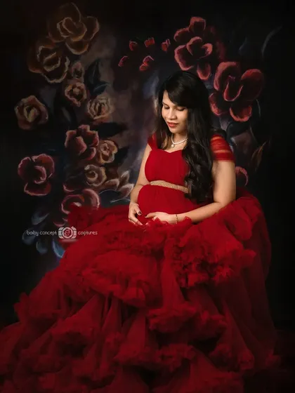 A seated pose from the red gown series, showing the mom-to-be cradling her bump. The voluminous red tulle spreads around her against the floral backdrop, creating a beautiful, painterly effect.