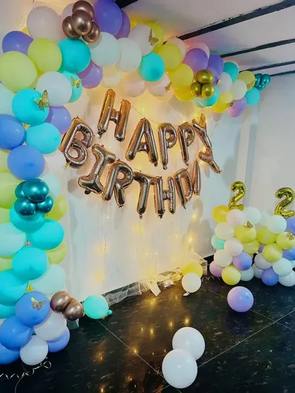 An indoor birthday party setup for a 22nd birthday. The decor features a colorful pastel balloon garland against a white curtain with fairy lights, creating a cheerful and pretty backdrop for photos.