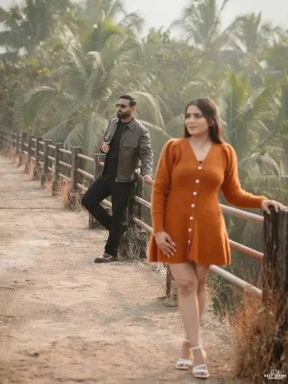 A stylish shot on a country road in Goa, with the bride in focus and the groom leaning against a fence in the background, creating a fashionable, layered portrait.