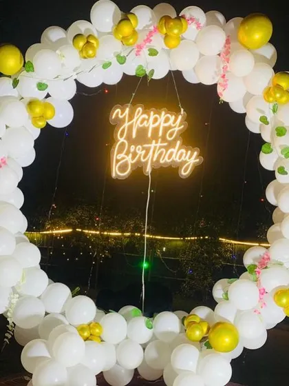 An outdoor birthday decoration featuring a full circular balloon ring in white and gold. The "Happy Birthday" neon sign at the center makes it a perfect backdrop for evening celebrations.