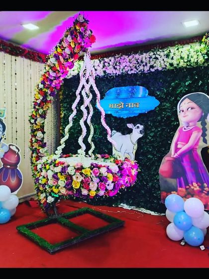 A Krishna-themed naming ceremony ('Namkaran') decoration with a floral hanging cradle. The green backdrop and character cutouts create a vibrant, traditional feel.