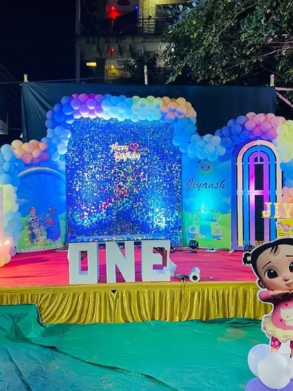 A wide view of an outdoor Cocomelon themed birthday stage, illuminated with blue lights and featuring a large 'ONE' sign and character cutouts.