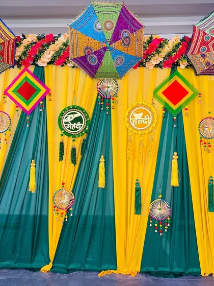 This is a classic fabric drape backdrop for a Haldi or Mehndi function. The combination of bright yellow and dark green curtains, accented with floral arrangements and traditional hangings, creates a vibrant and photogenic setting.