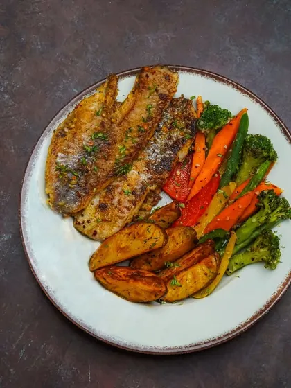 A grilled fish platter served with roasted potatoes and colorful vegetables. The moody lighting and rustic plate styling are perfect for a restaurant's social media feature.