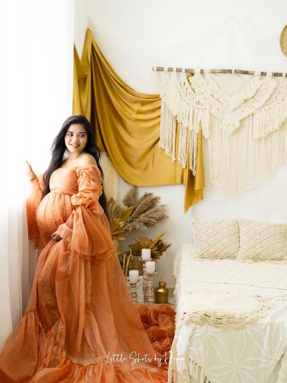 A beautiful portrait of a mother-to-be in a flowing peach gown. She stands by a window in a boho-chic room, looking out with a happy and serene expression.