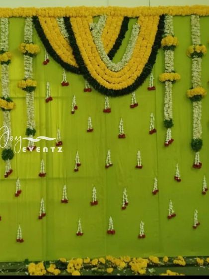 A simple and classic flower decoration for a half saree function. This backdrop uses a light green fabric base, adorned with hanging garlands of jasmine and marigolds in a traditional pattern.