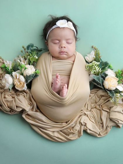 A simple and elegant floral arrangement. The baby is swaddled in a neutral wrap, surrounded by a bed of fabric and white flowers against a soft green background.