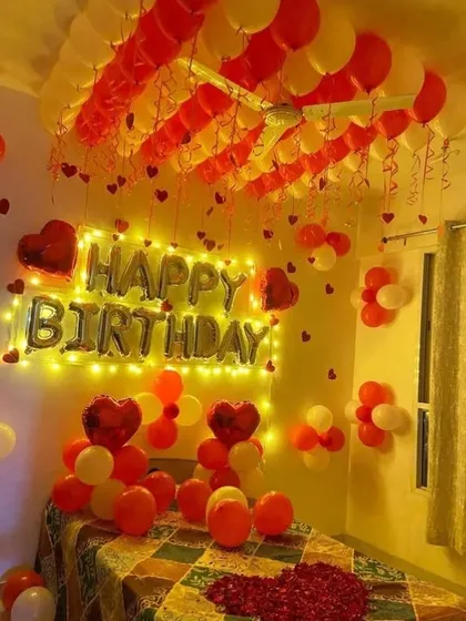 A cozy birthday room setup with red and yellow balloons. 'Happy Birthday' foil letters are lit by fairy lights, and the bed is decorated with rose petals.