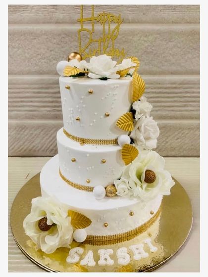 A grand and elegant three-tiered white cake, adorned with delicate piping, golden ribbons, and beautiful white and gold sugar flowers.