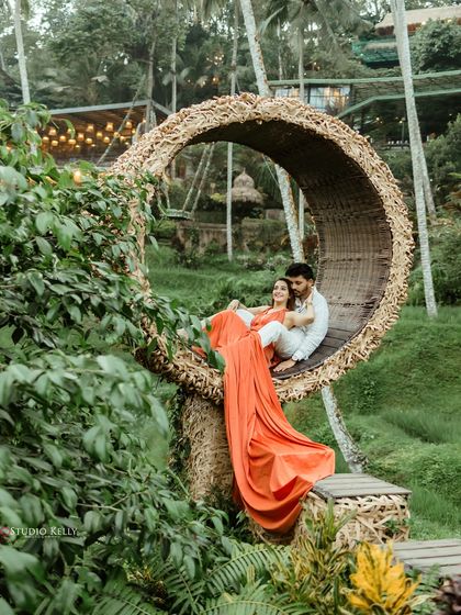 A romantic portrait of the couple nestled in a large, circular wicker seat amidst the lush rice paddies of Bali. The vibrant orange dress provides a beautiful pop of color.