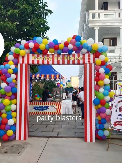The grand entrance to a carnival-themed party. A colorful balloon arch over a red-and-white striped gate welcomes guests, with personalized signs for the birthday kids.