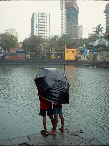 Two boys share an umbrella while standing at the edge of Banganga Tank. The modern high-rises in the background create a striking contrast with the ancient, sacred waters of the tank.