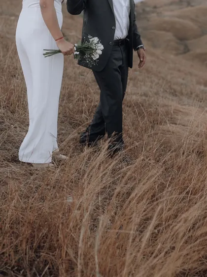 A detail shot focusing on the bride's elegant white jumpsuit and bouquet as she walks through the field, adding a touch of modern style to the natural setting.