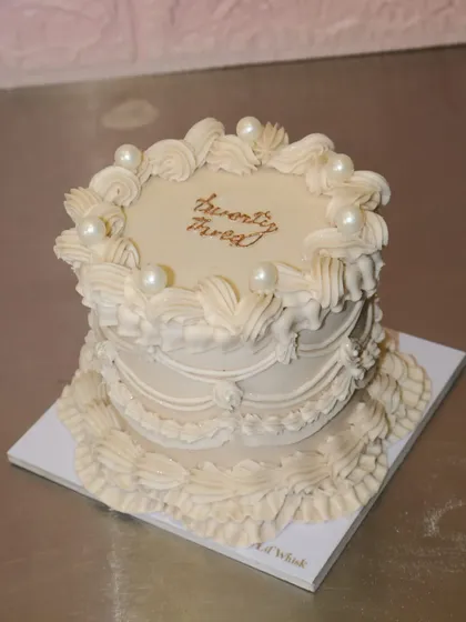 A beautifully decorated vintage-style cake in all white, featuring intricate piping and elegant pearl accents. The golden "twenty three" text adds a touch of class.
