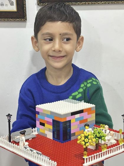 A proud young builder holds his colorful LEGO house, complete with a garden and lampposts.