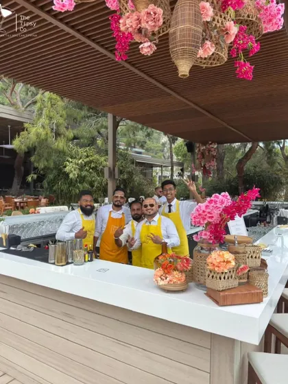 An outdoor bar setup designed for a tropical, daytime event. The white wood bar, floral arrangements, and rattan decor create a relaxed and stylish atmosphere, perfect for sipping cocktails by the sea.