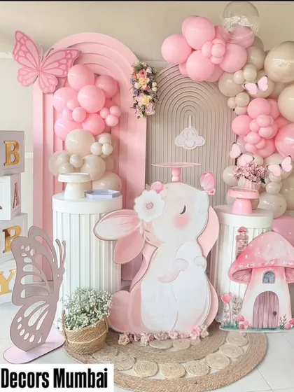 A sweet bunny and butterfly themed setup for a baby's party. The decor uses a soft pink color palette with props like a large bunny cutout, mushroom house, and "BABY" blocks.