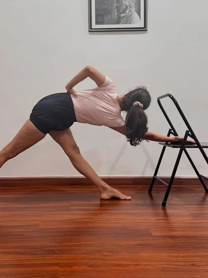 Parivrtta Trikonasana (Revolved Triangle Pose) is demonstrated here using a chair for support. The chair helps to maintain length in the spine and stability in the legs, allowing for a deeper and more refined twist.