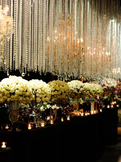 A long table at a cocktail event, lined with bouquets of white roses and illuminated by a curtain of hanging crystals and chandeliers.