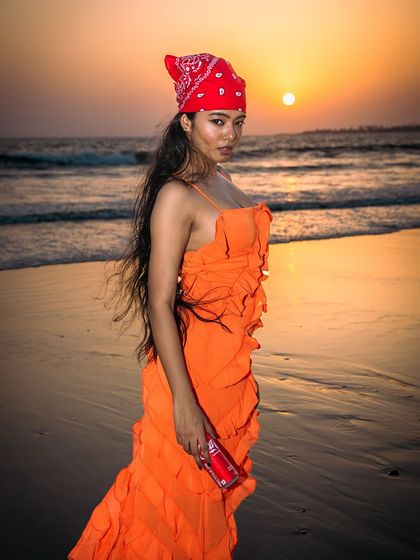 A vibrant sunset provides a stunning backdrop for this beach editorial. The styling with the orange dress and red bandana creates a bold, retro-inspired look.