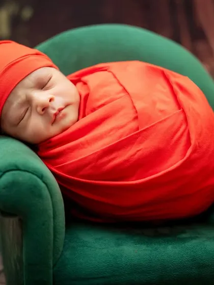A newborn wrapped in a red swaddle and sleepy hat rests on a miniature green sofa next to a crochet bunny.