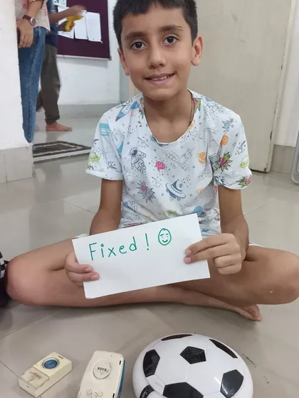 This young fixer is all smiles after successfully repairing a toy walkie talkie and a football. He is holding a "Fixed!" sign, a symbol of his newfound skills and confidence.