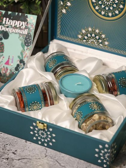 A luxurious teal-colored box with satin lining, filled with dry fruit jars and a candle. The intricate design on the jars adds to the premium feel of this Diwali hamper.