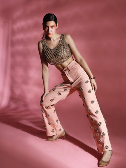 This look pairs intricately embellished pink trousers with a unique woven gold top. The dynamic pose and dramatic shadows highlight the textures and craftsmanship of both pieces.