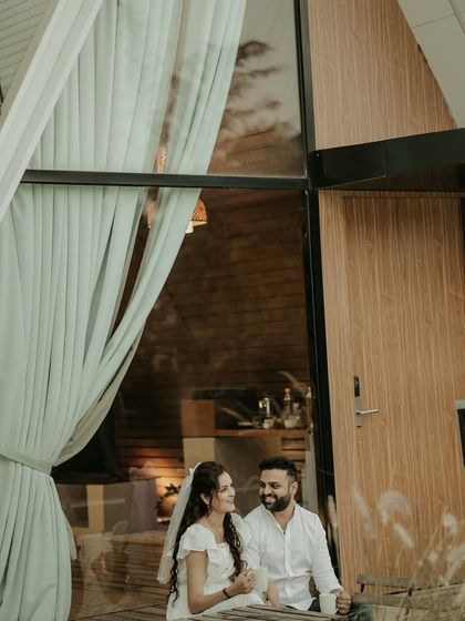 A quiet moment shared over coffee outside a modern A-frame cabin. The reflection in the large window and the soft curtains create a cozy and intimate pre-wedding scene.