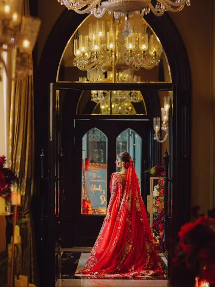 A breathtaking bridal portrait. The bride's magnificent red lehenga with its long train fills the frame as she stands in a grand, opulent doorway, looking like a queen.