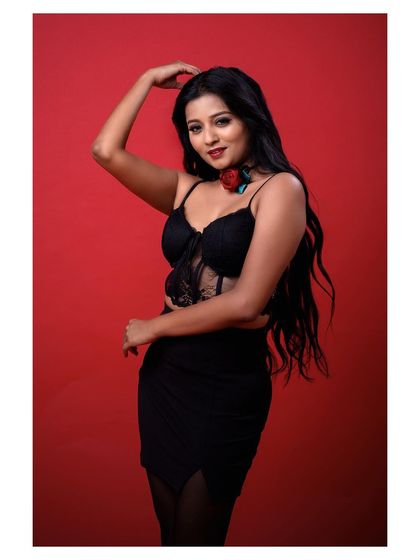 A full-body shot of a model in a glamorous black outfit. The pose is powerful and confident, set against a rich red background for maximum impact.