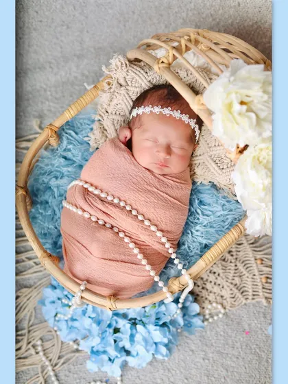 A newborn girl wrapped in a peach swaddle with pearls, sleeping peacefully in a round cane bassinet. The soft blue and white flowers provide a gentle, dreamy backdrop.