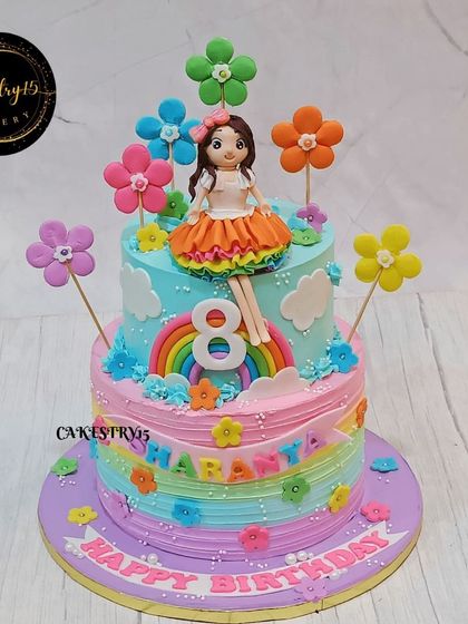 A full view of the delightful two-tier butterscotch cake for an 8th birthday. It's adorned with a doll topper, a fondant rainbow, and colorful flowers, creating a fun and vibrant look.