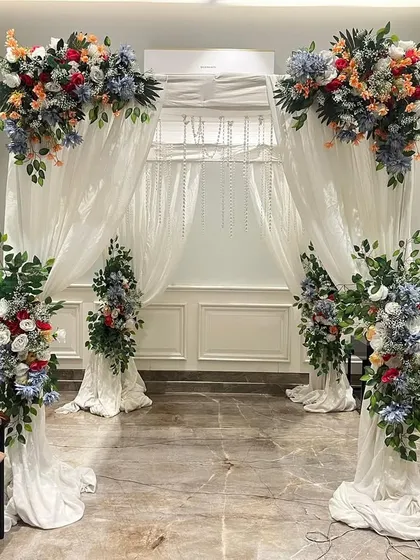 An elegant floral archway for a wedding or engagement ceremony. The setup uses white drapes and lush arrangements of artificial flowers to create a beautiful entrance.