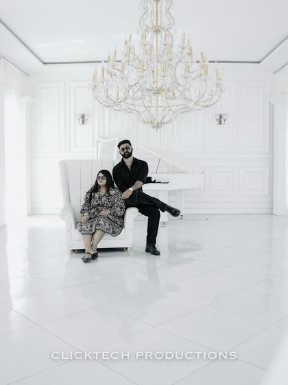 A wide-angle shot of a couple in a minimalist, all-white studio with a grand piano and chandelier, highlighting a clean, high-fashion aesthetic for their pre-wedding shoot.