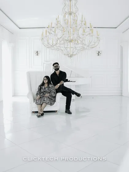 A wide-angle shot of a couple in a minimalist, all-white studio with a grand piano and chandelier, highlighting a clean, high-fashion aesthetic for their pre-wedding shoot.