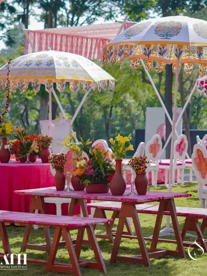 Guest seating at a vibrant outdoor event. The pink benches and tables are set under traditional block-printed umbrellas, surrounded by lush greenery.