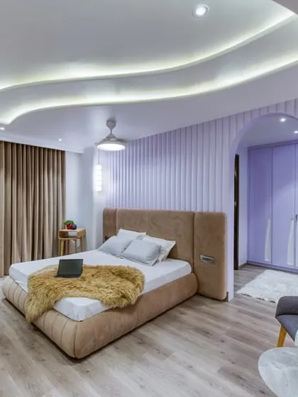 A playful yet sophisticated bedroom with a wavy, illuminated ceiling design and a fluted lavender accent wall. The arched doorway leads to a matching closet space.
