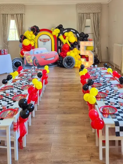 A complete room setup for a Disney Cars party. The long guest tables are decorated with checkered runners and themed tableware, all leading to the main backdrop with a giant Lightning McQueen.