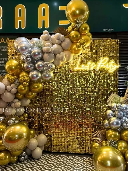 A stunning gold shimmer wall paired with a sophisticated balloon arch in gold, silver, and muted sand tones. The 'Lets Party' neon sign invites guests to celebrate in style.