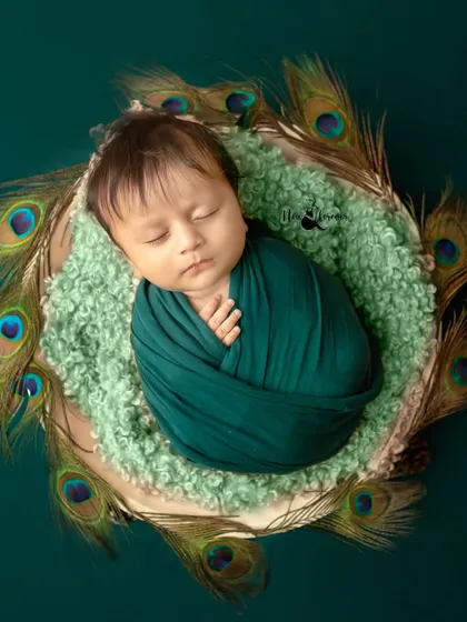 A top-down view of a newborn Krishna, nestled in a basket adorned with peacock feathers. The deep green wrap complements the theme perfectly.