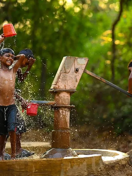 A young boy raises a red bucket, about to douse his friend, while another works the pump. This photograph captures the anticipation and action of their water game.