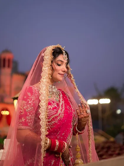 A beautiful North Indian bride in a pink lehenga, her veil framing her face perfectly against the evening sky.